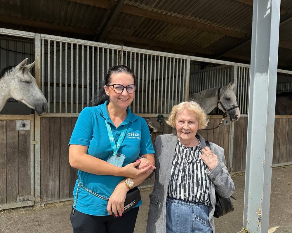 An Otter Care Professional and a client smiling together at a stables, with two horses looking over the doors behind them.