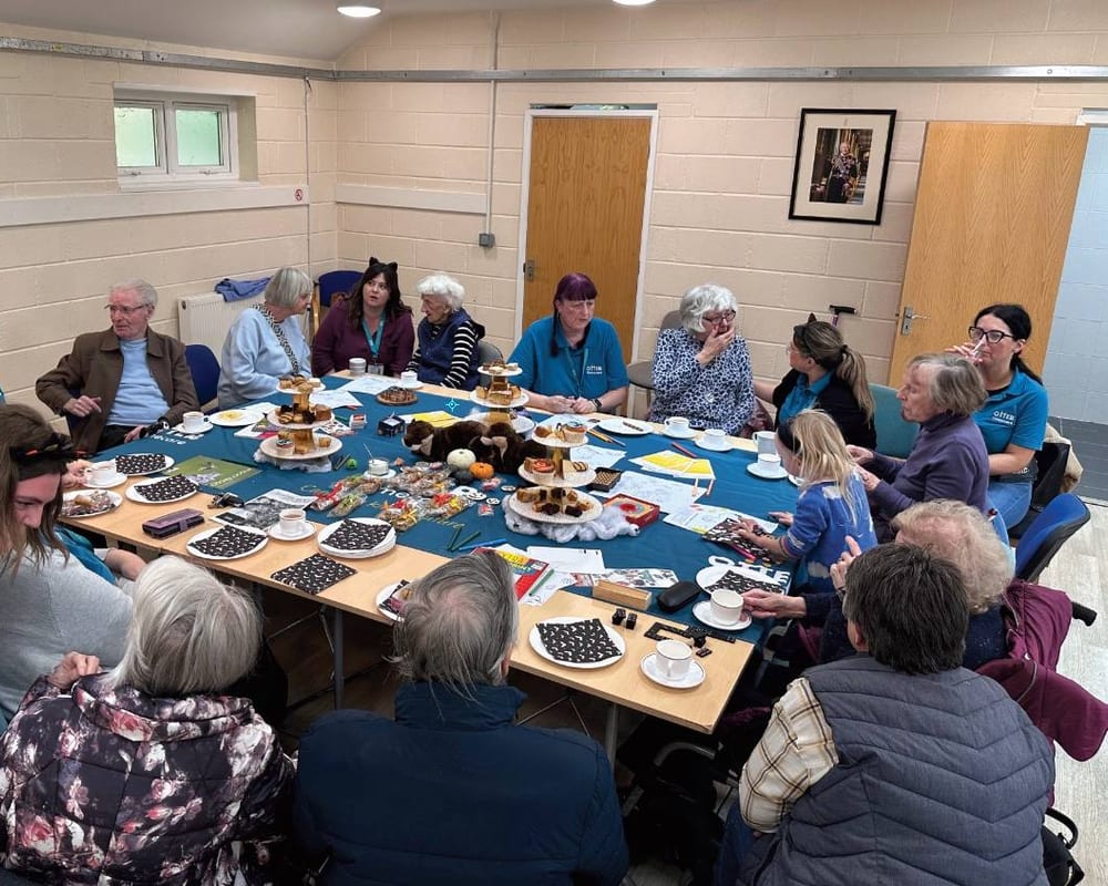 People gathered round a long table at an Otter Café session, tea and cake in front of them.